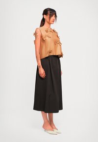 Ženska s tetovažami, oblečena v bež brezrokavnik s pentljicami, črne široke hlače skrajšanega kroja in bele spredaj konične slip-on petkice, stoji v profilu.