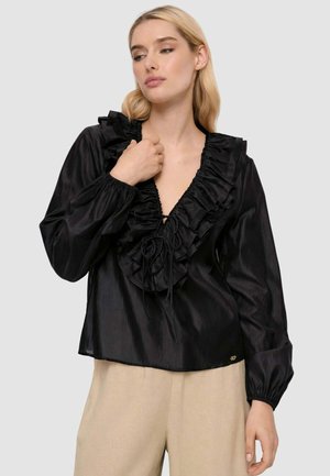 Donna che indossa una camicetta di satin nera con scollatura a V arricciata e maniche lunghe, abbinata a pantaloni beige, su uno sfondo grigio uniforme.