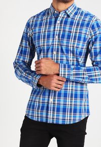 Uomo che indossa una camicia a quadri blu, bianca e nera con bottoni, mentre sistema il polsino della manica sinistra, con pantaloni neri.