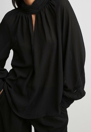 Løs sort tekstureret bluse med lange puffede ærmer og en høj, rynket halsudskæring med en lille V-åbning foran.