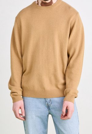 Mann mit Vitiligo, trägt einen beige-farbenen Rundhalspullover und hellblaue Jeans vor einem einfarbigen weißen Hintergrund, im Bereich von Hals und Oberschenkeln beschnitten.