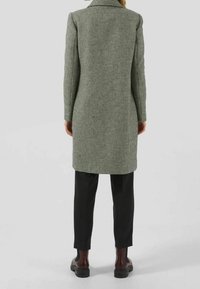 Abrigo largo con estampado de pata de gallo en tonos verdes y negros, que cuenta con un cuello y un corte recto, combinado con pantalones negros y botas robustas.