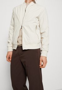 Jasnobeżowa kurtka bomberka wykonana z teksturowanego materiału, z zamkiem z przodu, bocznymi kieszeniami i ściągaczami przy rękawach. Nosi się ją na neutralnej bluzce i brązowych spodniach.