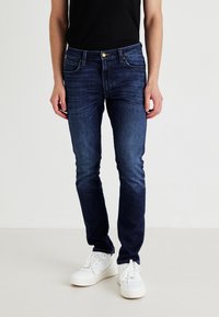 Jeans azul oscuro de denim, con un diseño clásico de cinco bolsillos, corte ajustado y un sutil desvanecimiento. Combinados con zapatillas blancas.