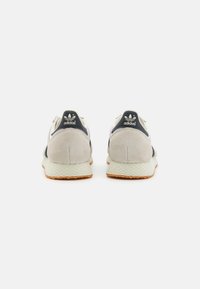 Adidas kedas gaiši pelēkā zamšā ar tumši zilām akcentiem un melno logo. Iekļauj teksturētu baltu zoli un jēlcukura gumijas ārējo zoli.