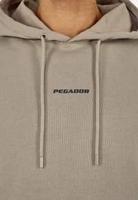 Felpa beige realizzata in morbido tessuto con cappuccio regolabile, con un logo scuro "PEGADOR" sul petto e terminali metallici sui lacci.
