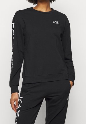 Person iført en sort EA7 sweatshirt og matchende sorte bukser med hvide EA7 Emporio Armani logoer på bryst, ærme og ben.