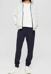 Lichtgekleurde pufferjack met capuchon, ritssluiting aan de voorkant en elastische manchetten; gecombineerd met marineblauwe joggingbroek, elastische broekspijpen en witte sneakers.