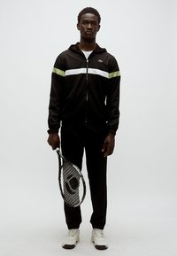 Musta urheilupuku, jossa valkoiset ja limenvihreät raidat, seisova mieshahmo, joka pitelee tennismailaa ja on pukeutunut valkoisiin lenkkareihin.