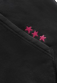 Czarna tkanina o gładkiej fakturze z trzema różowymi haftowanymi gwiazdkami w pobliżu kieszeni, podkreślającymi detale projektu.