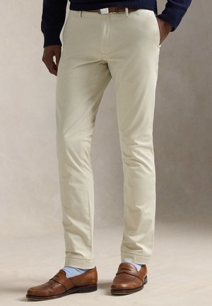 Ember bézs színű, keskeny szabású nadrágot, barna bőr mokaszint, világoskék zoknit, tengerészkék pulóvert és barna övet visel. Kezei a zsebében vannak.