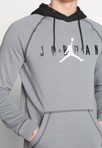 Sudadera gris con detalles en negro, bolsillo canguro, puños de canalé y logo "JORDAN" impreso en blanco y negro. Cuenta con costuras planas.