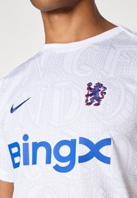 Hvit fotballtrøye med et subtilt tekstmønster, blå "Bingx"-logo og brodert blå løvekappe. Laget av lett, pustende materiale.