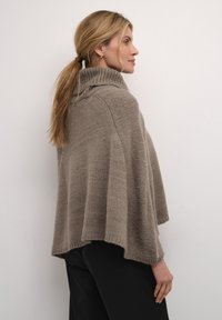 Beez värvi kudum ponšo kõrge kaeluse ja laienevate varrukatega. Pehme tekstuur, vabalt langev lõige ja soonikkoes servad. Kantud koos tumedate pükstega, näidatud küljelt.