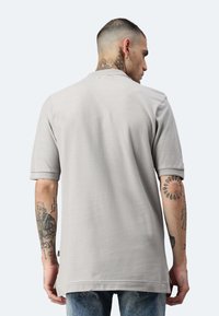 Camisetta grigia a maniche corte con una texture a coste, caratterizzata da una vestibilità ampia e un orlo arrotondato. Tatuaggi visibili sulle braccia, con uno stile casual.