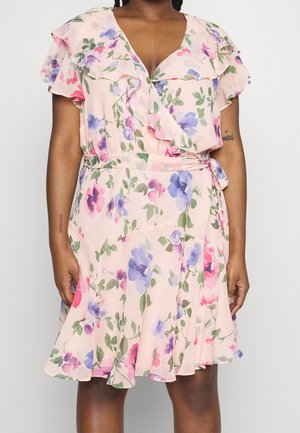 Femme portant une robe portefeuille rose à fleurs, avec des manches volantées et une ceinture à la taille, ornée de fleurs violettes, bleues et roses accompagnées de feuilles vertes.
