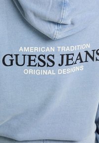 Gaiši zils hoodijs ar regulējamu kapuci; melnā un baltā krāsā drukāta teksta uzraksts "American Tradition Guess Jeans Original Designs."