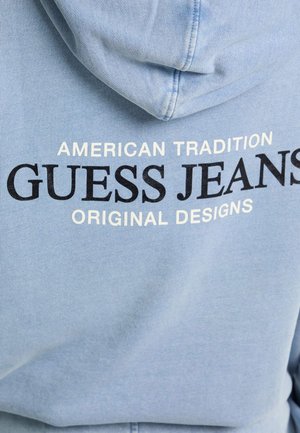 Jasnoniebieska bluza z kapturem z sznurkiem; nadrukowany tekst w czerni i bieli brzmiący "American Tradition Guess Jeans Original Designs."