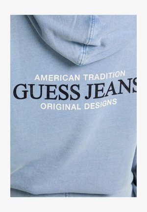 Jasnoniebieska bluza z kapturem z sznurkiem; nadrukowany tekst w czerni i bieli brzmiący "American Tradition Guess Jeans Original Designs."