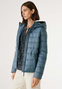 Vrouw in een blauwe gewatteerde capuchonjas over een donkere gebloemde blouse en lichtgekleurde broek, die naar de zijkant kijkt tegen een effen achtergrond.