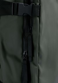 Primer plano de una mochila de tela verde, con una cremallera de calidad, correa ajustable y un cierre resistente con acabado mate.