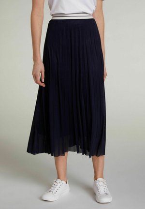 Mujer que lleva una falda midi plisada azul marino con una cinturilla blanca, combinada con zapatillas blancas y una camisa blanca metida por dentro.
