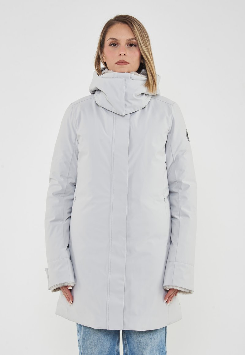 Donna che indossa un cappotto invernale grigio chiaro con cappuccio e foderato in pelliccia, in piedi contro uno sfondo bianco uniforme, rivolta verso l'avanti.