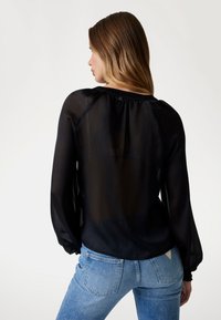 Svart genomskinlig blus med långa puffiga ärmar, veckad textur och knäppning bak, tillsammans med ljusblå denimjeans.