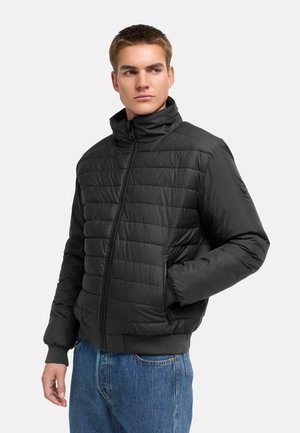Ung mand iført en sort quiltet jakke med lynlås og blå jeans, stående med den ene hånd i lommen og kigger til siden.