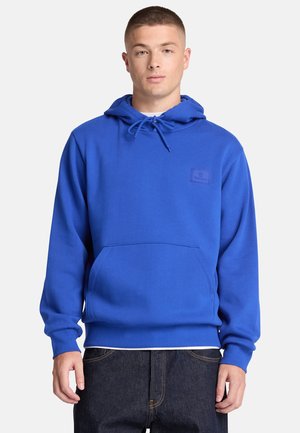 Jonge man met kort haar die een blauwe hoodie met voorzak en een donkere spijkerbroek draagt, staand tegen een effen witte achtergrond.