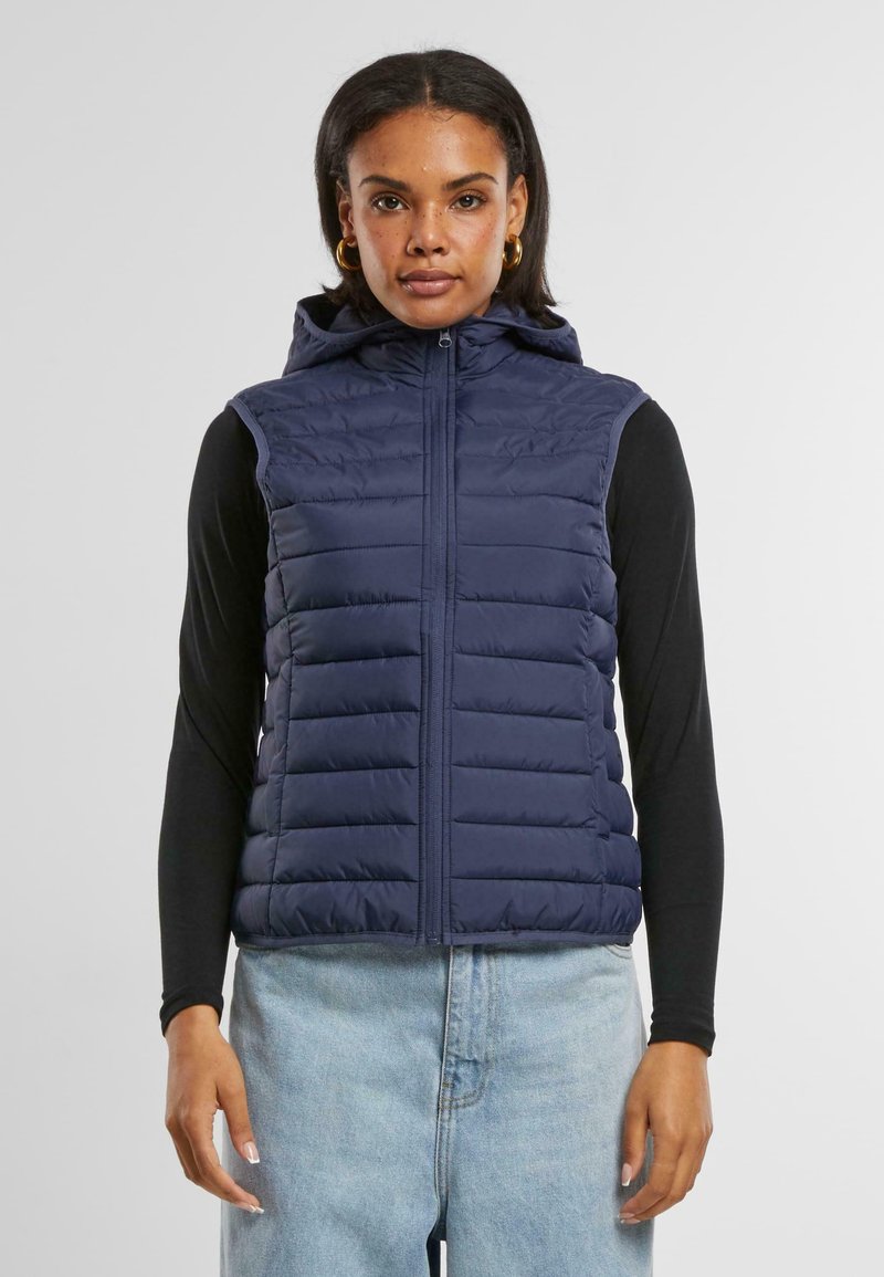 Gilet bleu marine rembourré avec une fermeture éclair devant et une capuche. Présente un design matelassé et des poches latérales. Porté par-dessus un shirt à manches longues noir.