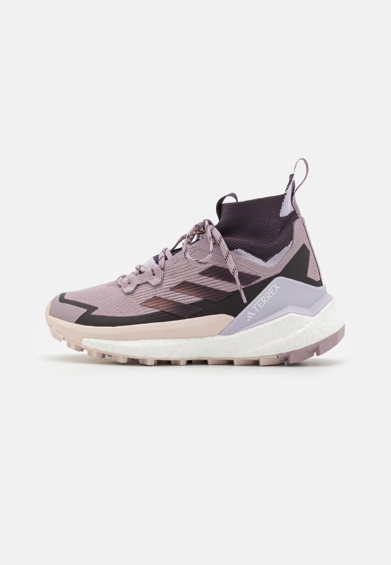 Svijetlopurpurna atletska cipela s teksturiranim mrežastim gornjim dijelom, crnim detaljima i podstavljenim bijelim potplatom. Ima visoki ovratnik i reflektivne detalje.