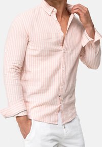 Mann trägt ein hellrosa und weiß gestreiftes Hemd mit hochgescrollten Ärmeln und weißen Hosen, Hand die Kragen anpasst.