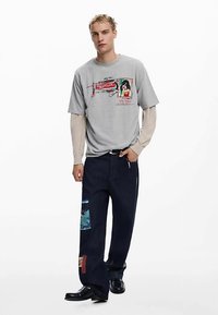 Tricou grafic gri cu imprimeuri, purtat peste un tricou cu mâneci lungi bej. Pantaloni din denim întunecat cu pete și croială dreaptă; pantofi negri.