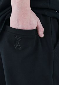 Black cotton sweatpants with a soft texture, featuring a side pocket and an embroidered dark 'K' logo on the front.