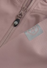 Ljust rosa, texturerad tygjacka med en svart dragkedja och en silverfärgad logotyp. Designen är stilren med minimal detaljer.