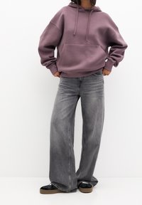 Felpa viola oversize con tasca frontale, abbinata a jeans grigi a vita alta e larghi. L'outfit include scarpe nere con suola beige.