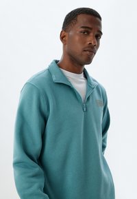Teal zip-up sweatshirt med hög krage, med en diskret logotyp på bröstet och textil med struktur. Vit skjorta under.