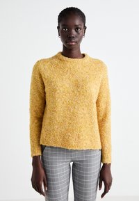 Un pull en tricot jaune avec une surface texturée et duveteuse, doté d'un col arrondi, associé à un pantalon à carreaux gris. La coupe est décontractée et informelle.