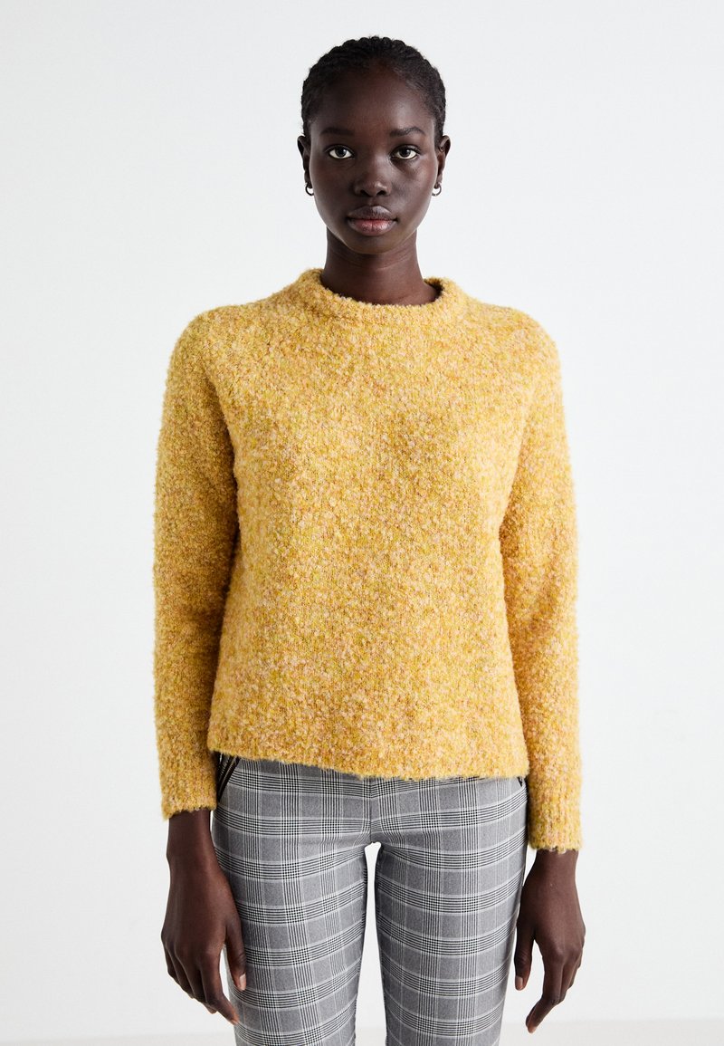 Un pull en tricot jaune avec une surface texturée et duveteuse, doté d'un col arrondi, associé à un pantalon à carreaux gris. La coupe est décontractée et informelle.