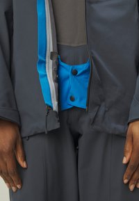 Fodera interna blu di una giacca scura, con un pannello grigio, caratterizzata da una tasca con bottone a pressione. Il materiale sembra impermeabile e liscio.