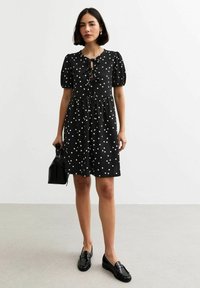 Abito nero con pois bianchi, maniche a palloncino corte e dettaglio di fiocco davanti. Collo rotondo e silhouette svasata. Indossato con mocassini neri.
