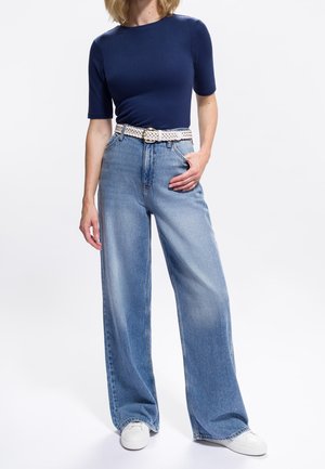 Marineblauw getailleerd T-shirt met korte mouwen, gecombineerd met lichtblauwe jeans met wijde pijpen, vastgehouden met een witte gevlochten riem en witte sneakers.