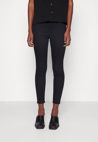 Personne portant un pantalon noir ajusté, des chaussures noires à enfiler à bout carré, et une chemise noire boutonnée, debout devant un fond blanc uni.