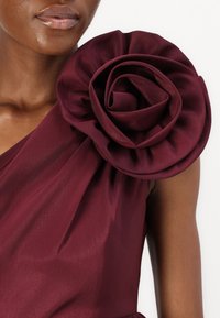 Burgundy-mekko, jossa on suuri kangasruusu olkapäällä. Sileä pinta ja laskostettu kangas luovat pehmeän siluetin.