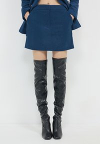Une personne portant une jupe en velours côtelé bleue et des bottes en cuir noires jusqu'au genou, se tenant devant un fond blanc uni.