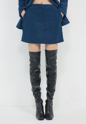 Person, die einen blauen Cordrock und schwarze kniehohe Lederstiefel trägt, steht vor einem schlichten weißen Hintergrund.