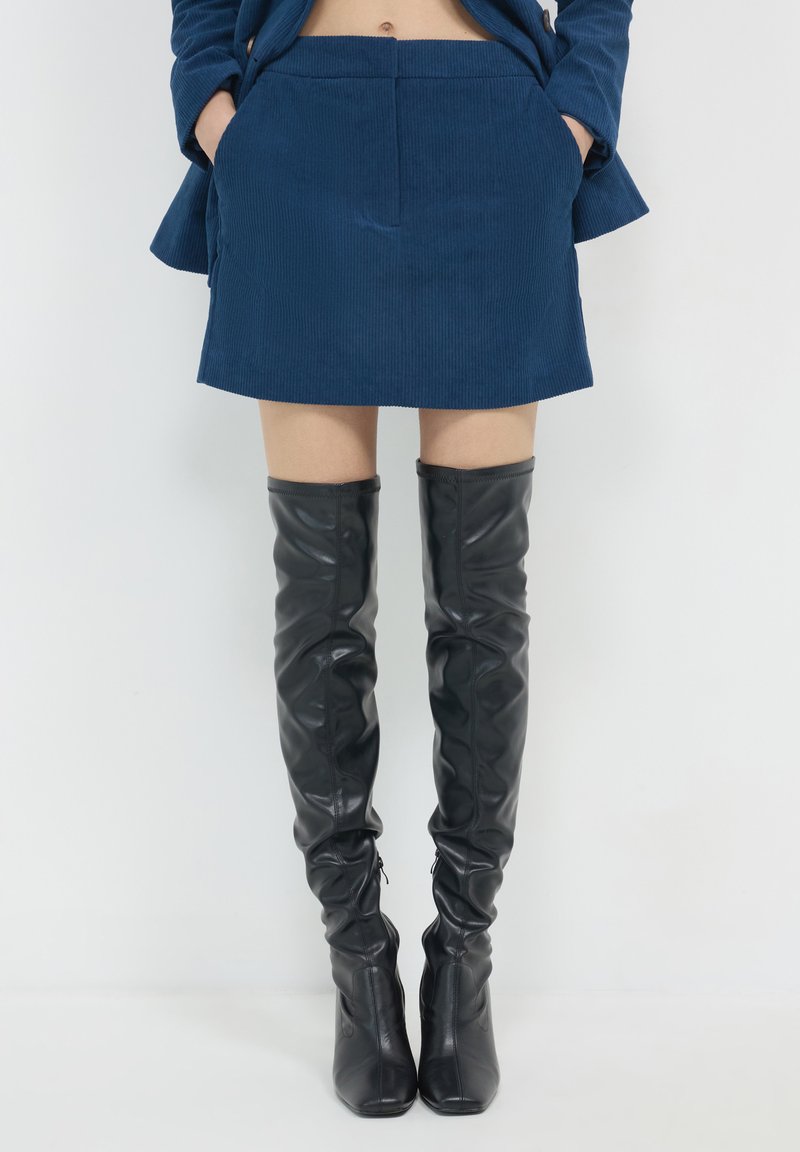 Une personne portant une jupe en velours côtelé bleue et des bottes en cuir noires jusqu'au genou, se tenant devant un fond blanc uni.