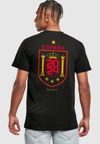 Fekete pamut póló, nagy piros és sárga Spanyolország címkével, arany koronával, "ESPAÑA 2024" felirattal és díszítő elemekkel alatta.