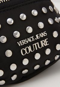 Černý látkový pouch s kovovými hroty a metalickým logem "Versace Jeans Couture". Uzávěr na zip, hladká textura, kompaktní design.